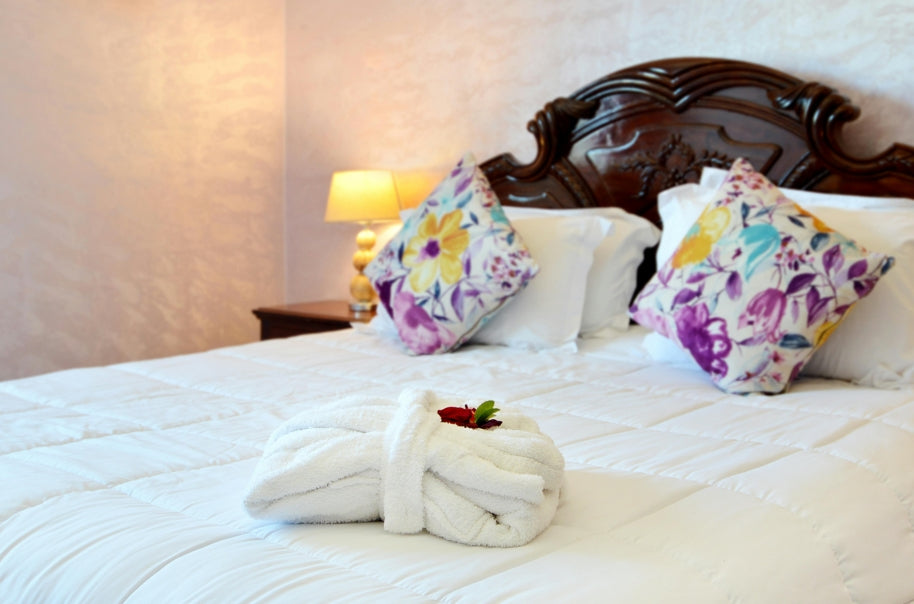 Luxurious hotel bed with white linens, floral accent pillows, and a plush folded bathrobe. High-end bedroom interior featuring a carved wooden headboard and warm bedside lighting, perfect for a relaxing stay or boutique travel experience.